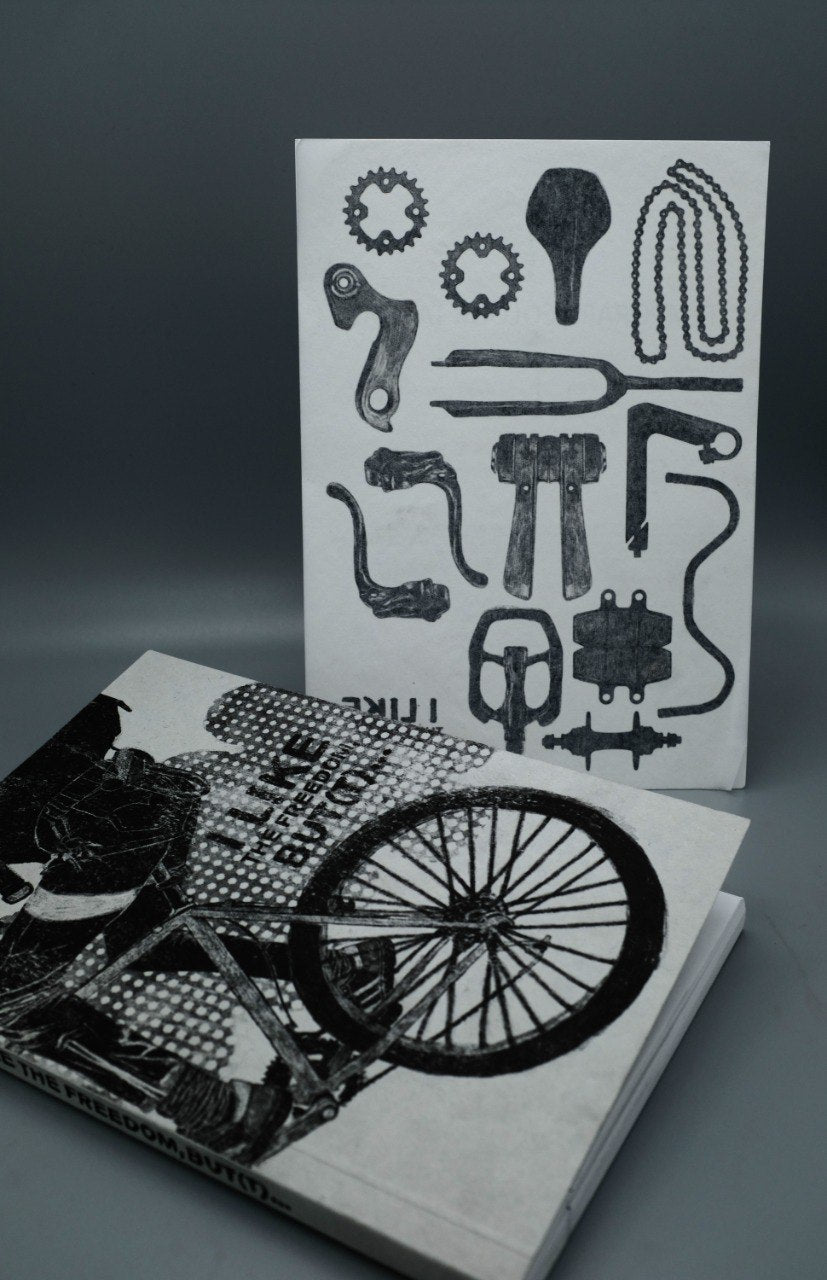 Product photos of the book "I like the freedom but(t)" laying on the floor, behind it, a sheet of temporary tattoos with bike parts on it.